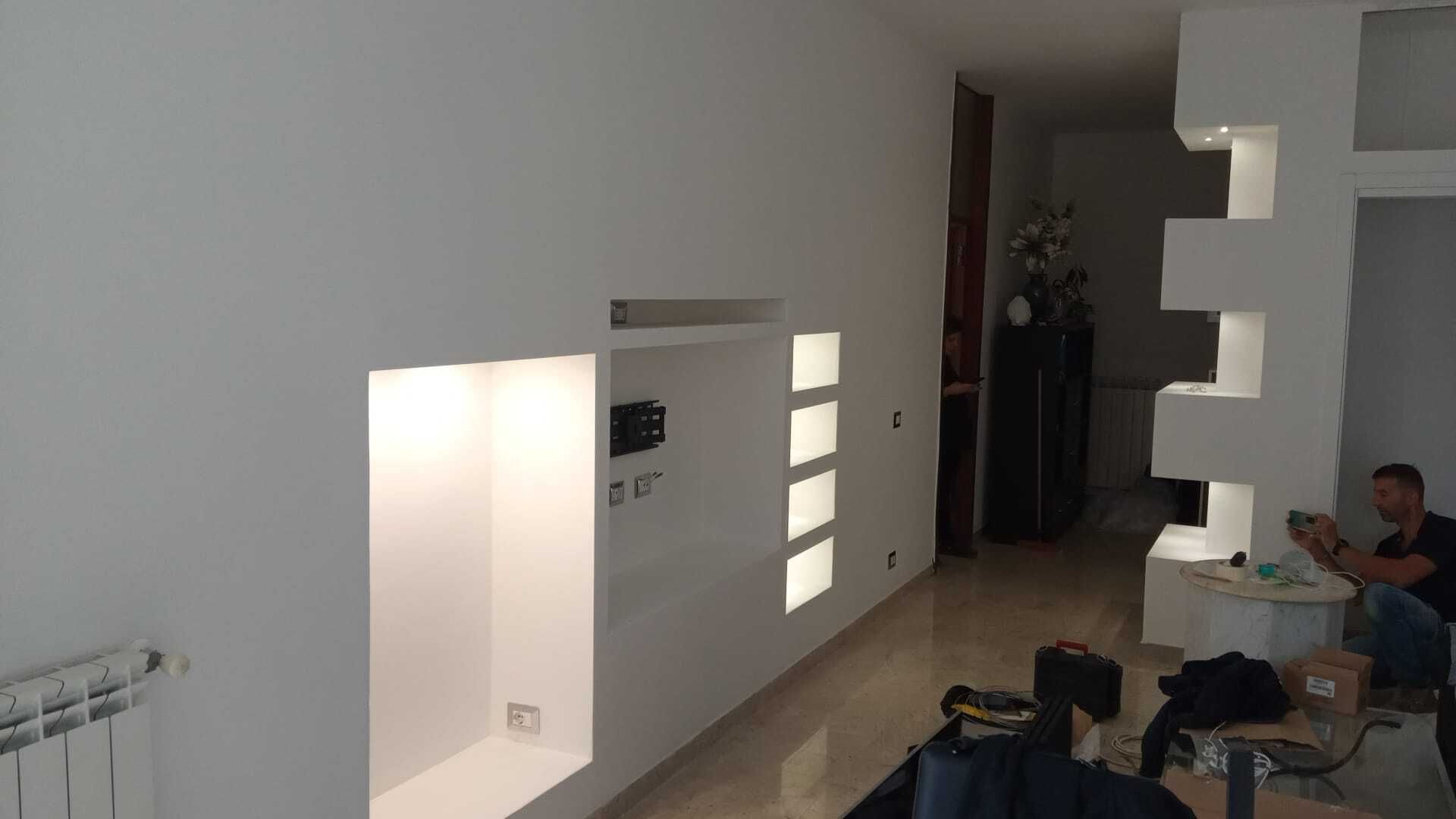 Soggiorno minimalista con mobili bianchi, mensole a parete e una persona seduta sul divano che usa un laptop.
