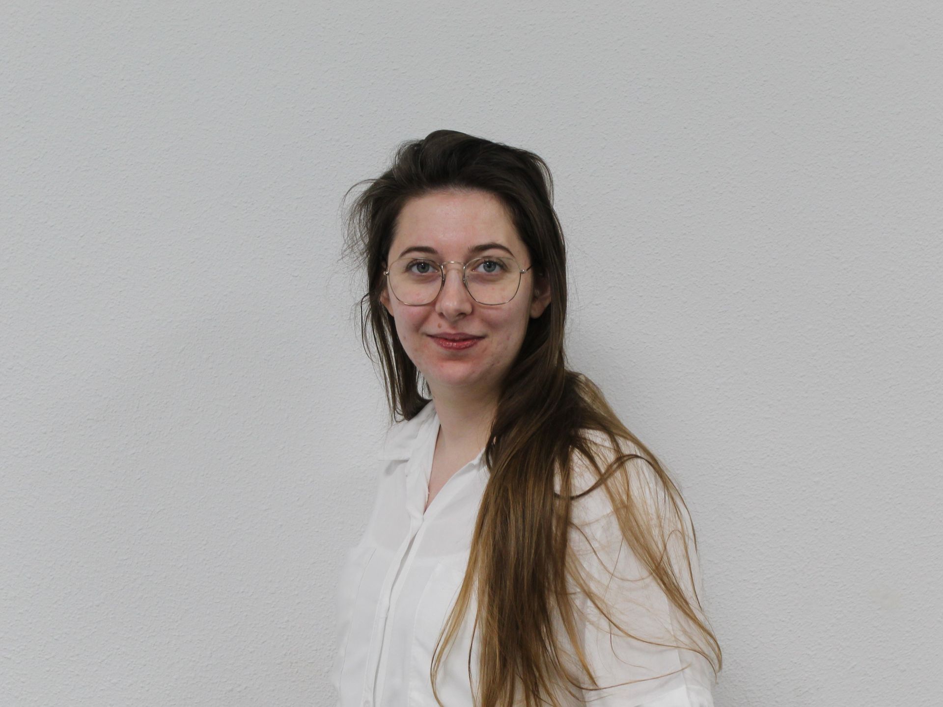 Een vrouw met een bril en een wit shirt staat voor een witte muur.