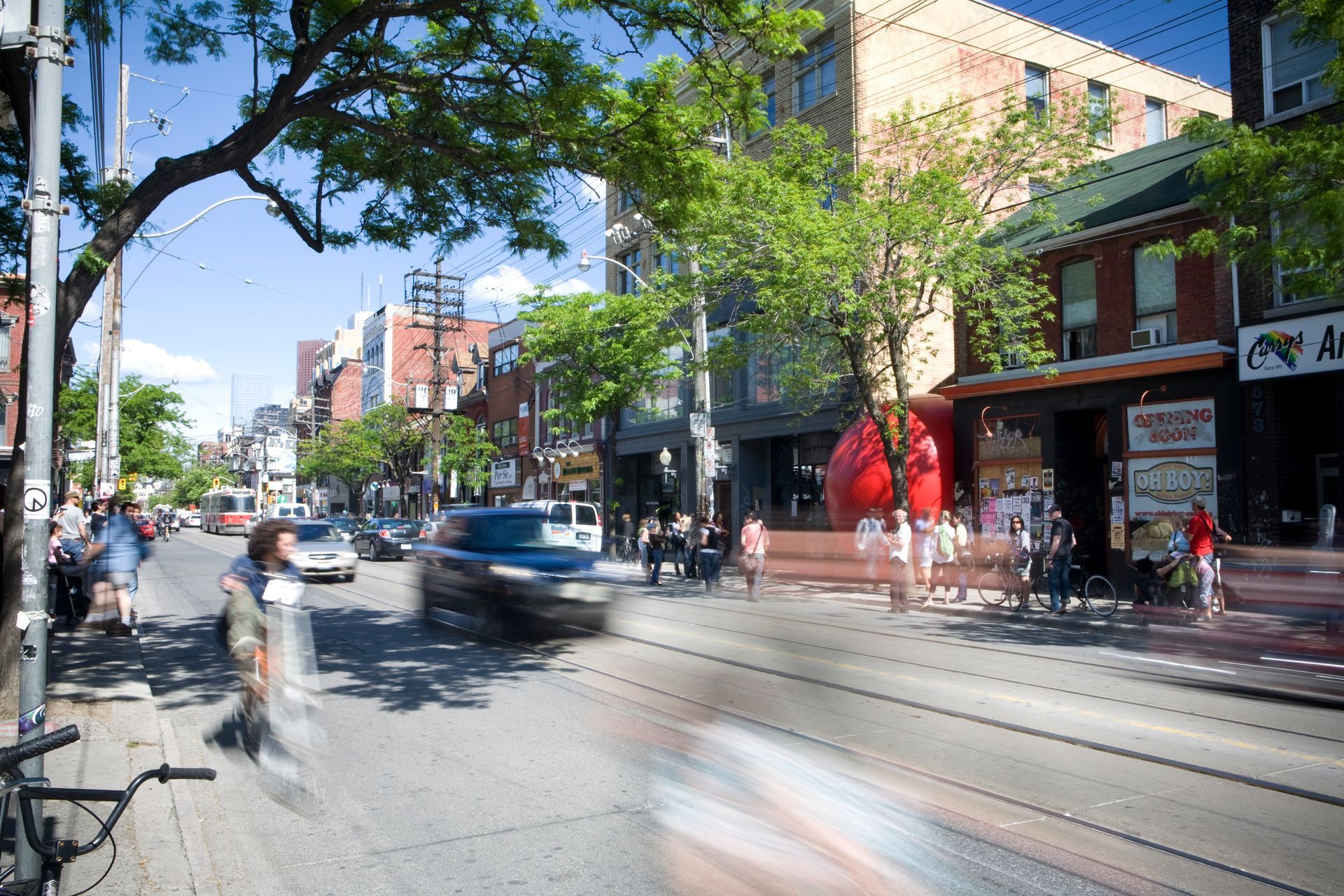 Street scene with blurred motion of cars and pedestrians; buildings, trees, and blue sky. Redball sits in an alleyway.