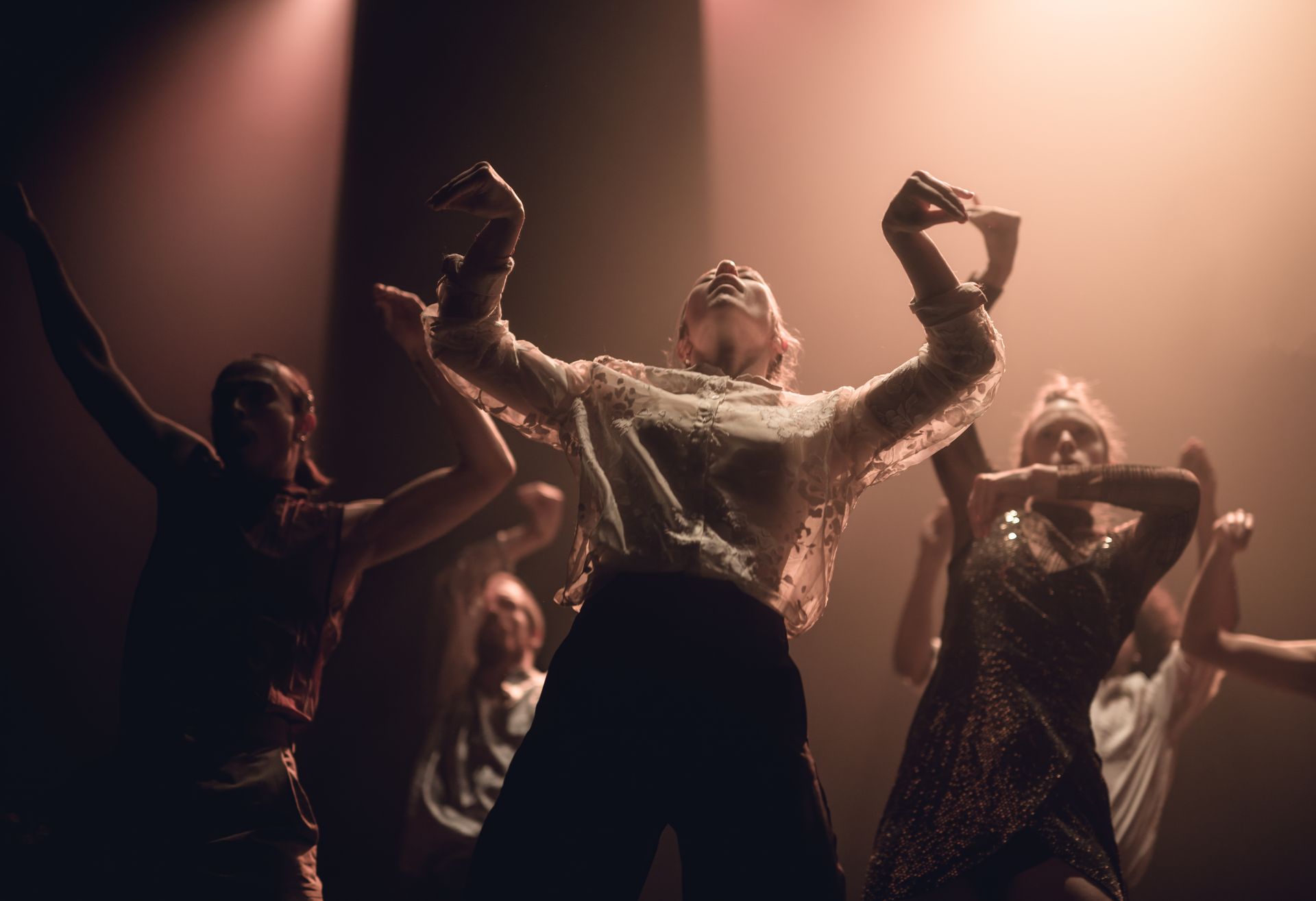Dancers in dim lighting, arms raised, mid-performance. Warm hues and silhouettes create a dramatic effect.
