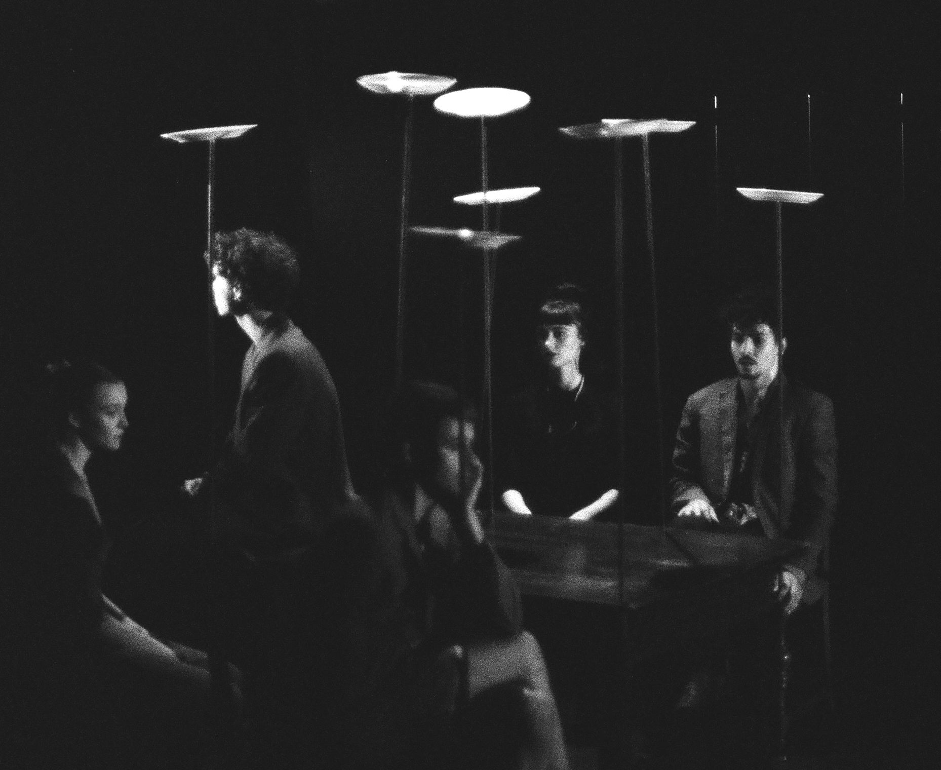 Five circus artists in formal clothes sit in black and white. Plates balancing on sticks surround them.