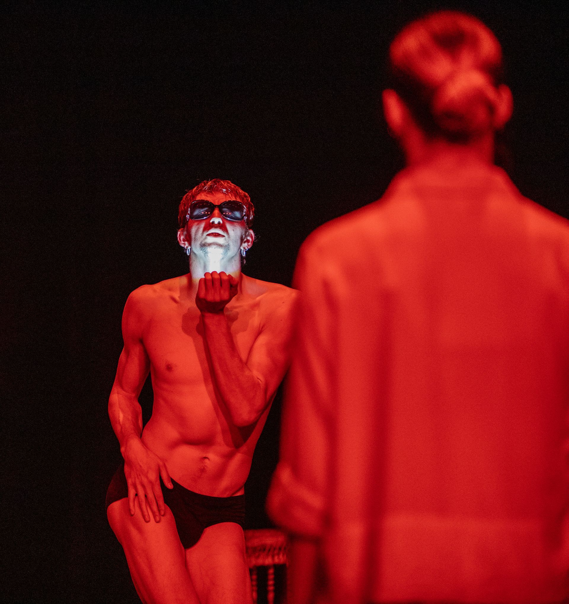 Man in sunglasses blows a kiss, lit dramatically with red light. Another person is in soft focus.