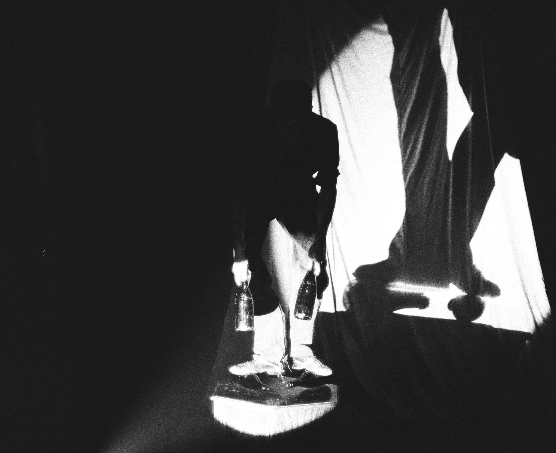 A shadowy figure holds glass bottles by their necks. Lit dramatically in black and white. 
