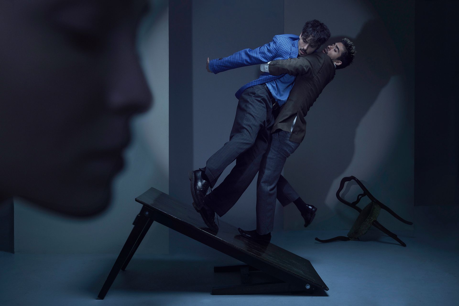 Two circus artist lean against each other on a broken tabletop. Another face in soft focus.