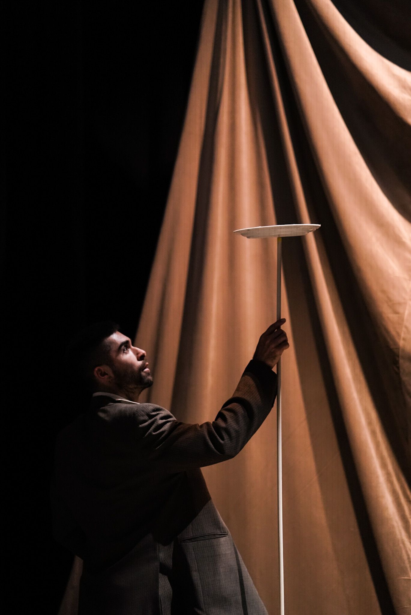 Circus artist in a suit balances a plate on a stick in front of draped fabric.