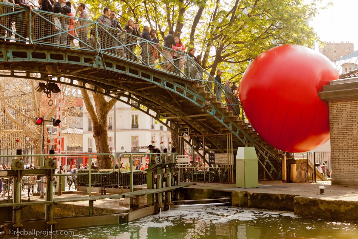 People stand on a bridge looking at a large red inflatable ball below