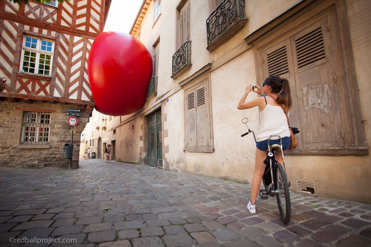 A person sitting on a bike takes a picture of a large red inflatable ball suspended in an alley