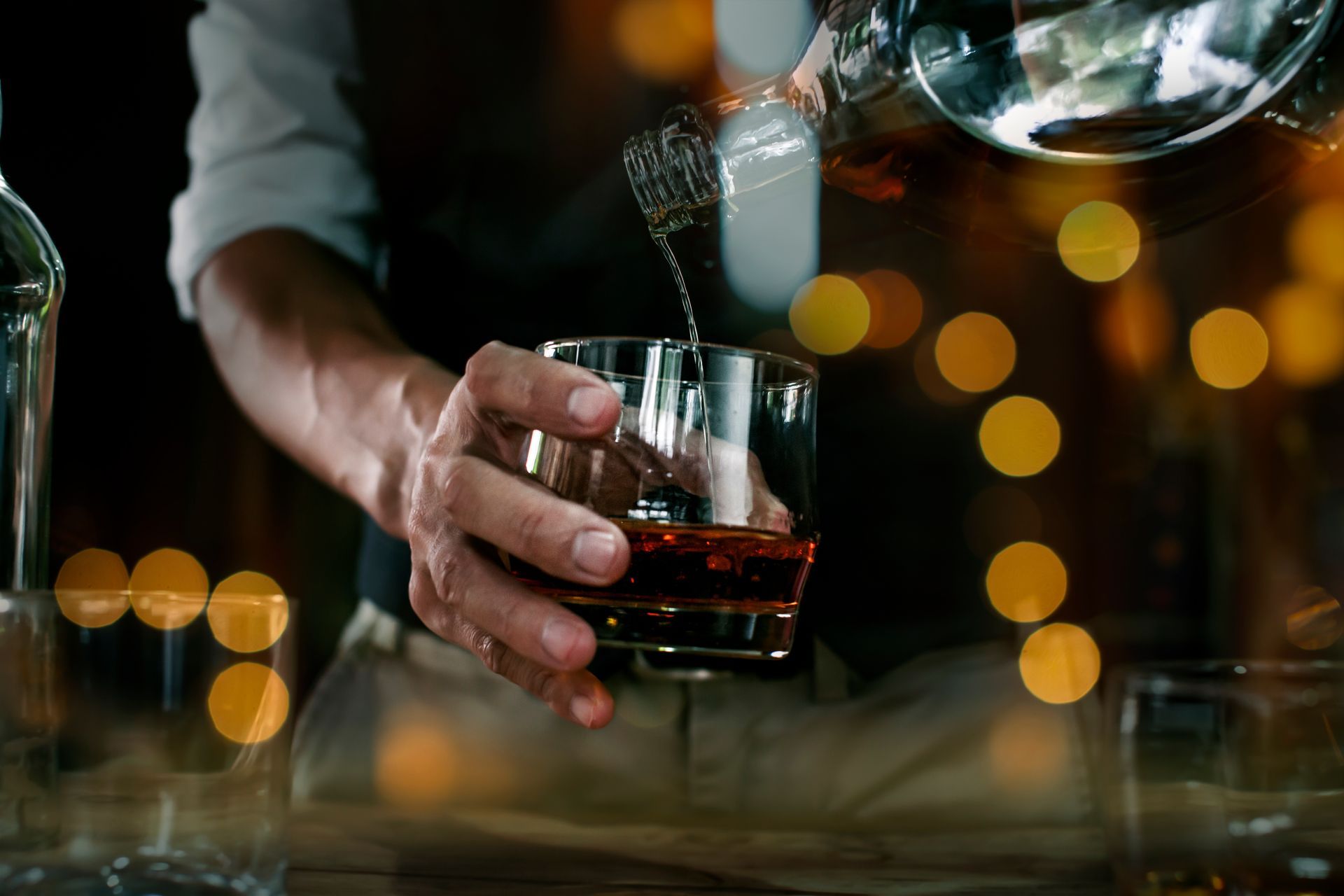 Hand pouring whiskey into a glass with warm bokeh lights in the background Hand pouring whiskey into a glass with warm bokeh lights in the background