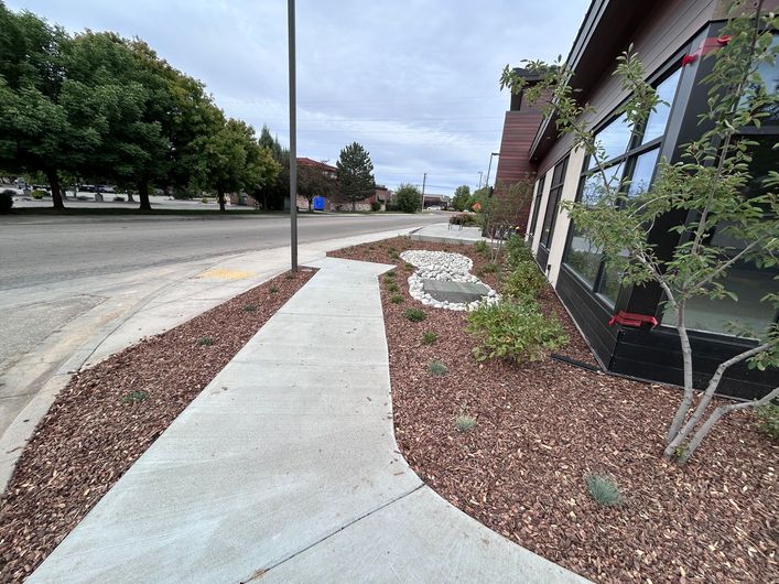 Sidewalk and landscaped path beside a building, with mulch, shrubs, and a parking lot beyond.