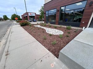 Sidewalk beside a brick storefront with large windows and a rock-mulched landscaped bed.