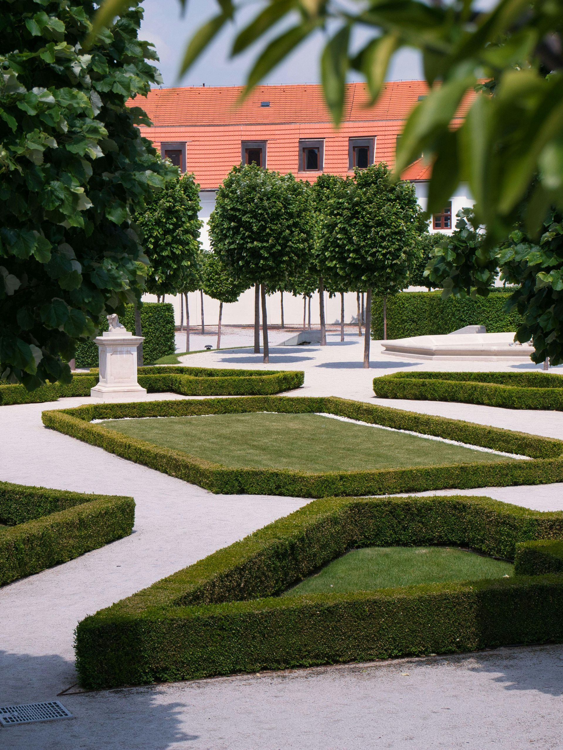Formal garden with geometric trimmed hedges and gravel paths, framed by green trees and a red-tiled building.