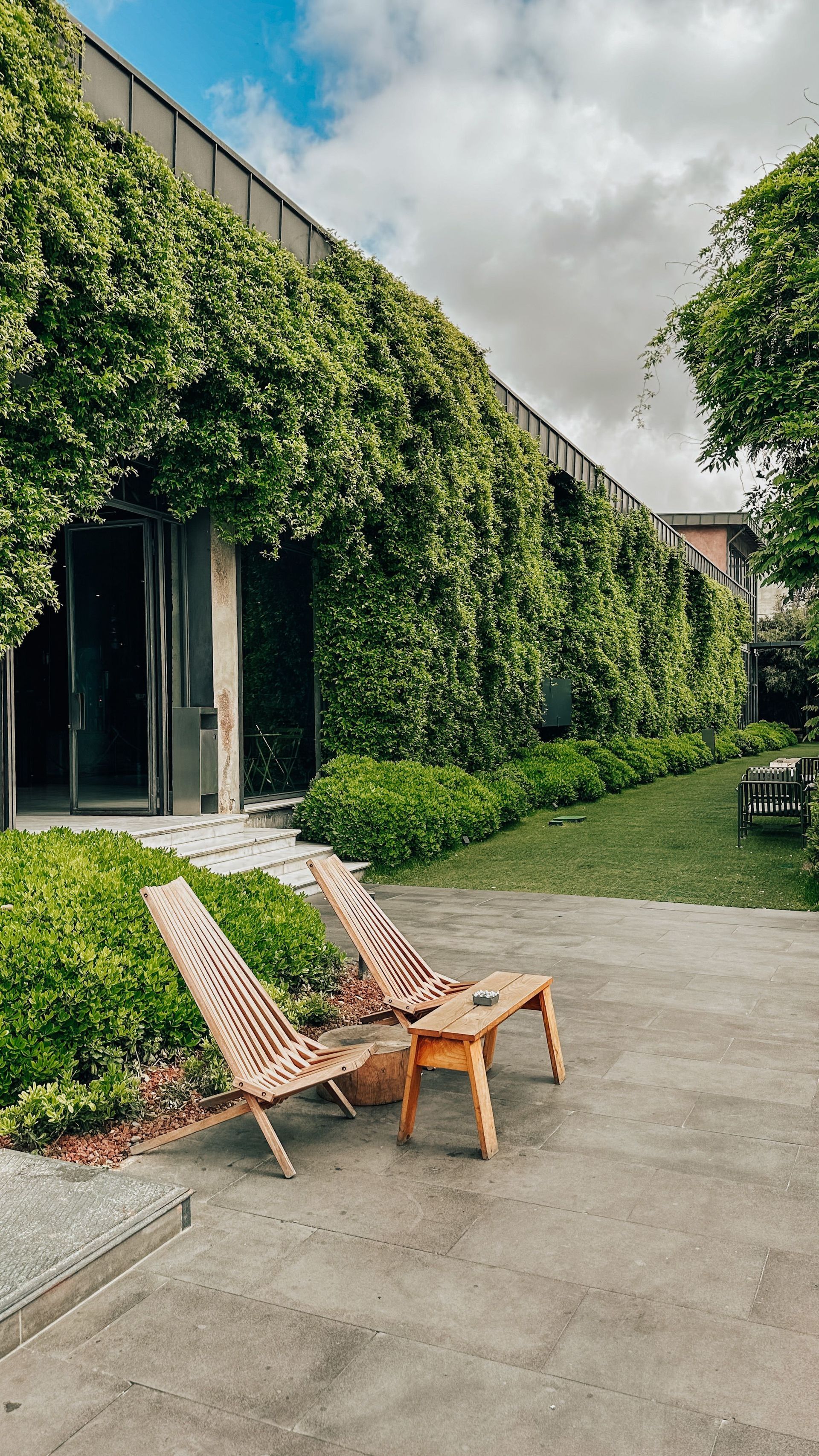 Two woven lounge chairs on a patio beside an ivy-covered building under a cloudy sky