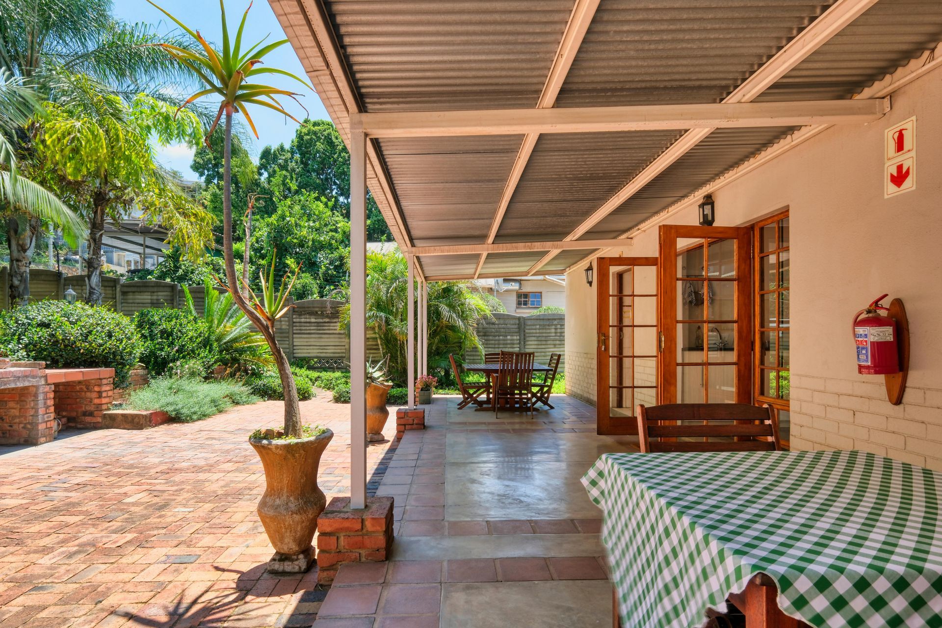 Covered patio with checkered tablecloth, open wooden doors, and a garden path in sunlight
