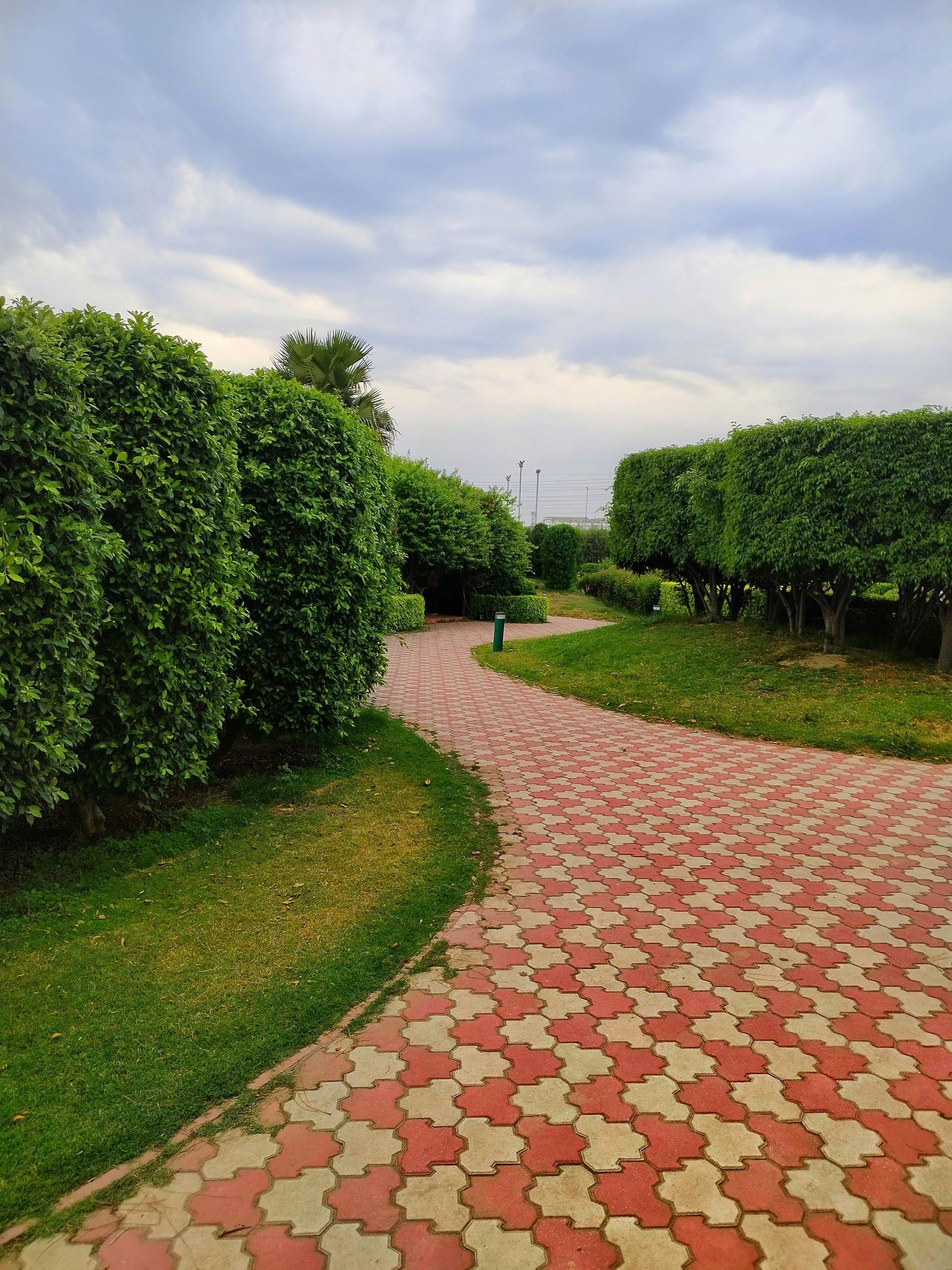 Curved red-and-white brick path through green hedges under a cloudy sky