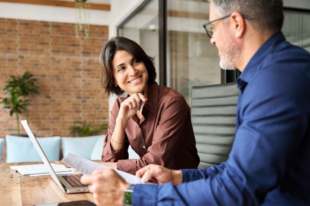 A man and a woman are sitting at a table in front of a laptop engaged in conversation