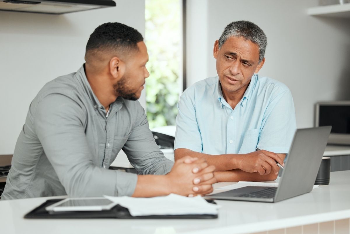 Two men are sitting at a table in conversation in front of a laptop computer.