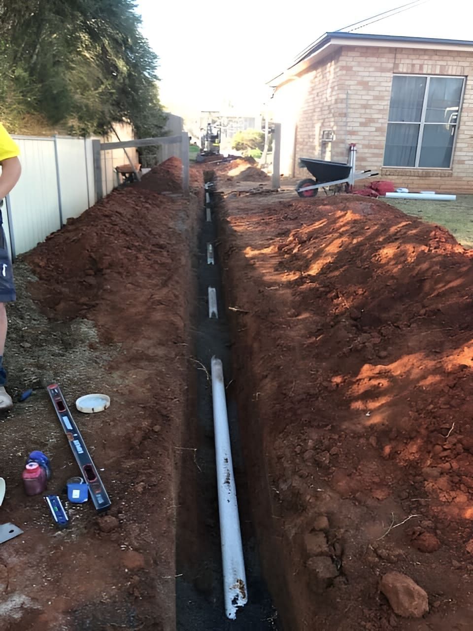 A Man Is Measuring A Pipe In The Dirt In Front Of A House — Nudges Plumbing In Dubbo, NSW