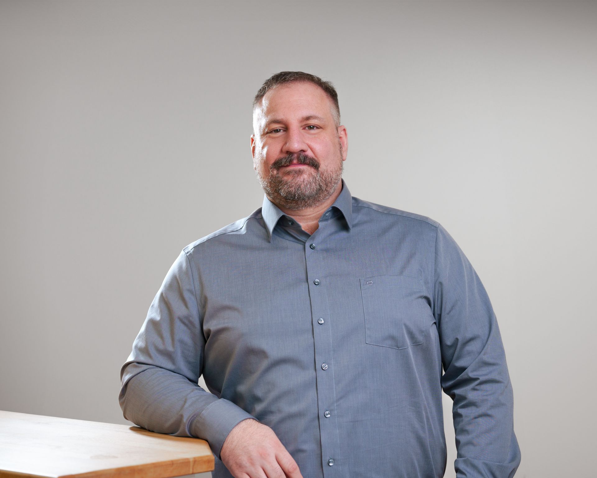 A man with a beard is standing next to a wooden table.