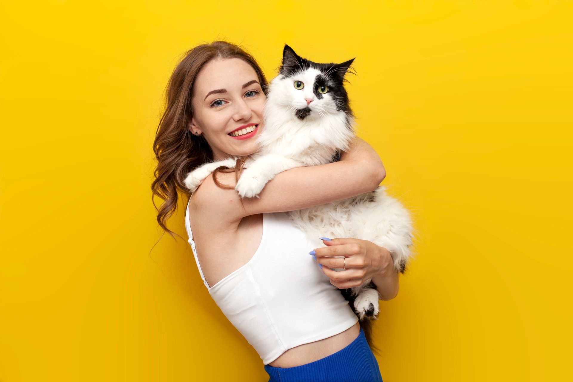 Woman in white top holding a fluffy black-and-white cat against a bright yellow background