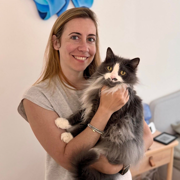 Woman holding a fluffy black-and-white cat indoors, both looking at the camera and smiling