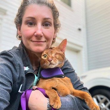 Woman holding an orange cat in a purple harness outdoors in front of a white house