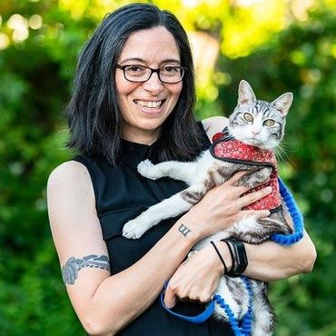 Woman with glasses holding a gray cat in a red harness outdoors, both facing the camera.