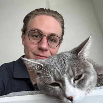 Person with glasses posing behind a sleepy gray cat indoors