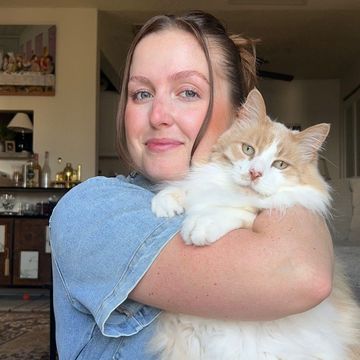 Woman in a blue shirt holding a fluffy orange-and-white cat indoors, both looking at the camera.