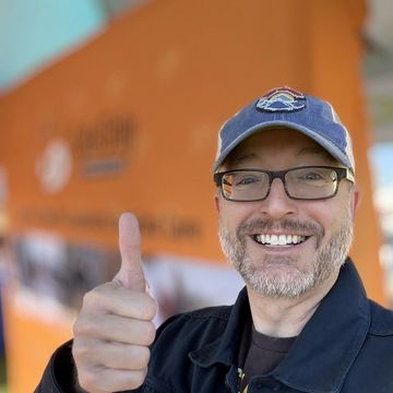 Smiling person in glasses and cap giving a thumbs-up in front of an orange truck