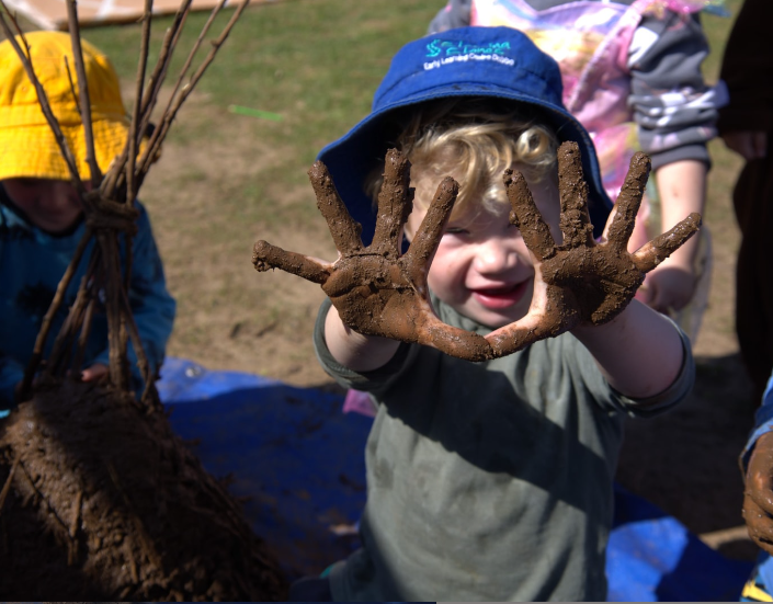 A young boy wearing a blue hat shows off his muddy hands