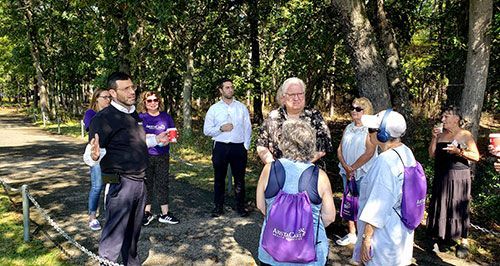 Group of people listening to a person with a beard gesturing in a wooded area, trees in the background.
