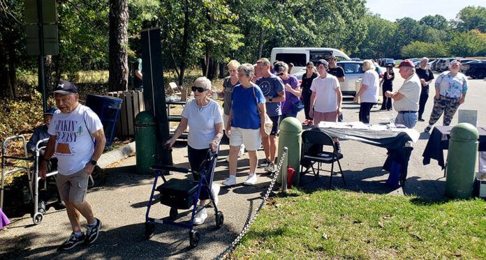 People gathered outdoors; some with walkers.  Several standing near a table. Sunny day; trees and parked cars in background.