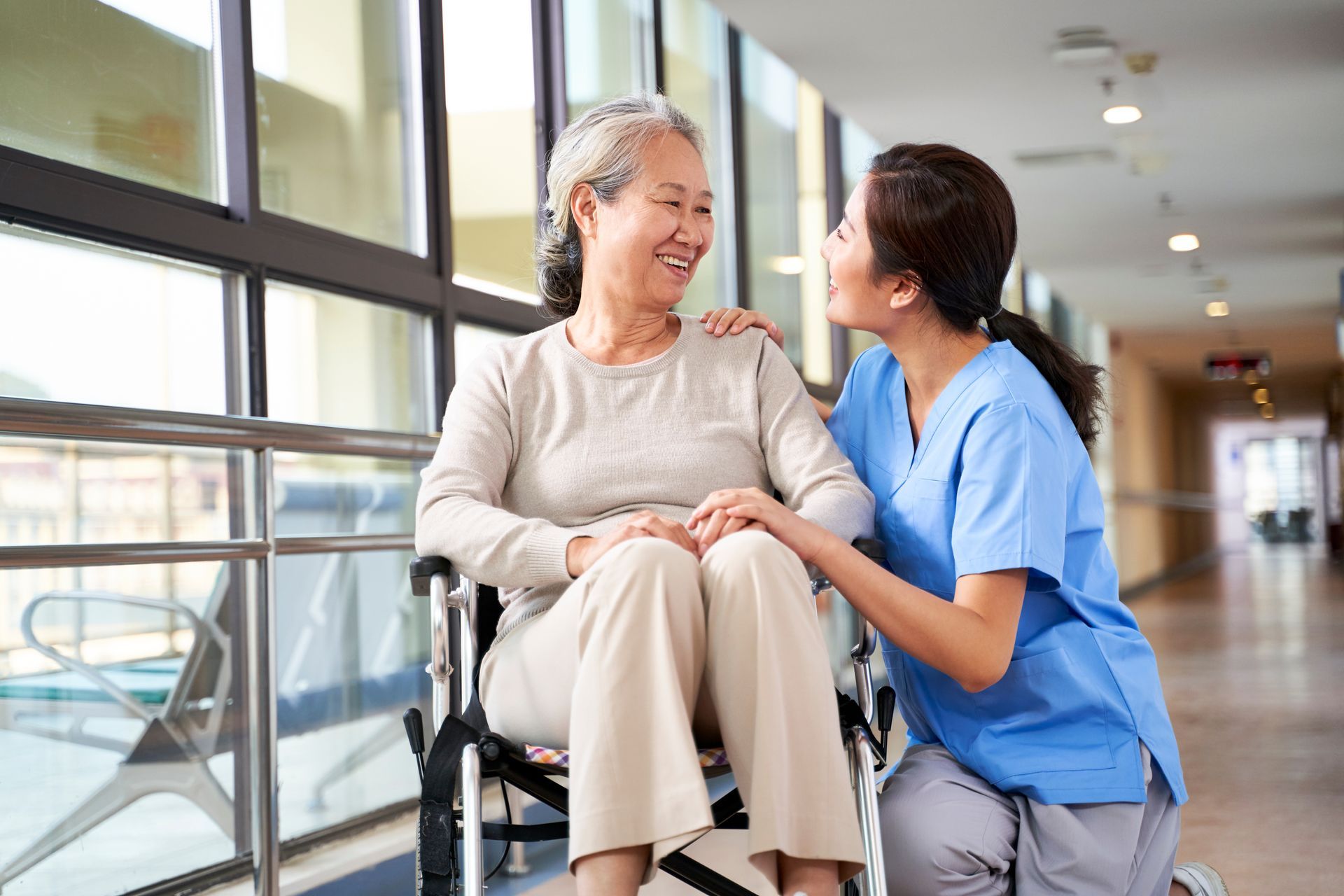 A nurse is kneeling down next to an elderly woman in a wheelchair.