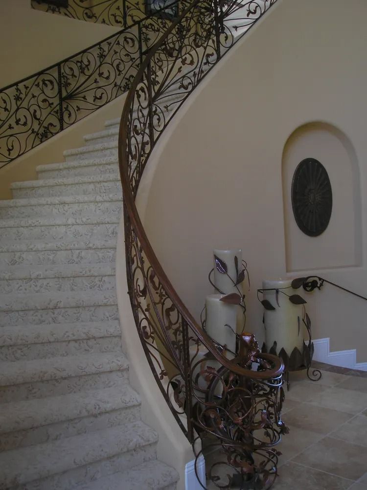 Elegant curved staircase with ornate wrought iron railing, beige walls, and decorative pottery.