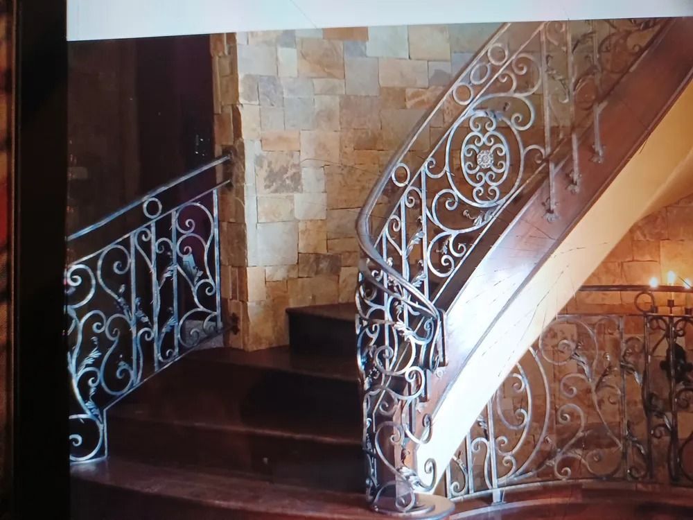 Ornate wrought iron railing on a wooden staircase with stone wall backdrop.