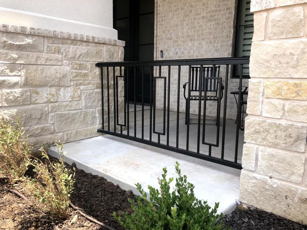 Black metal railing on a concrete patio with stone walls, next to landscaping.