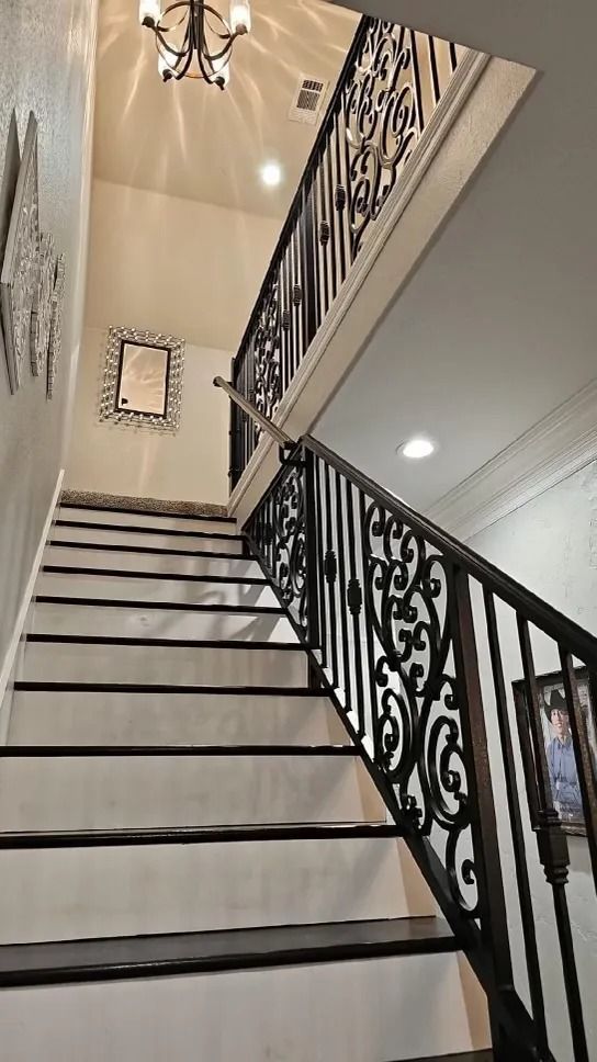 Staircase with black treads, ornate wrought iron railing, white walls, and a decorative chandelier.