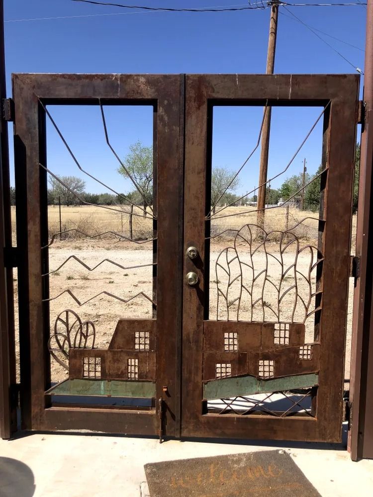 Rustic metal double gate with cut-out design of a house, trees, and landscape, set against a distant field and blue sky.