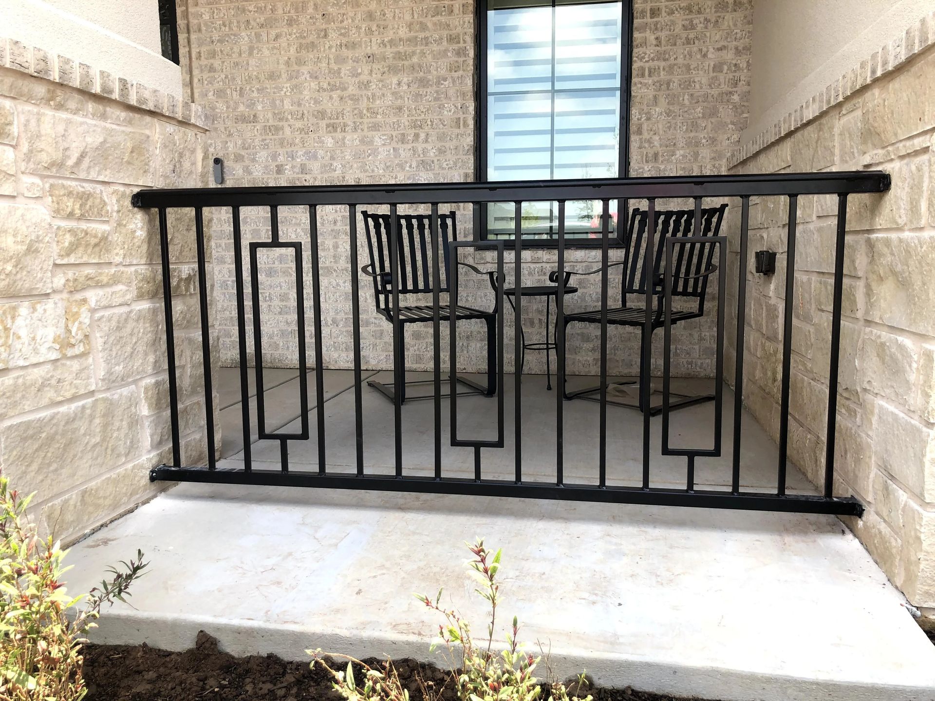 Black metal porch railing frames a small patio with a table and chairs. Beige brick wall.
