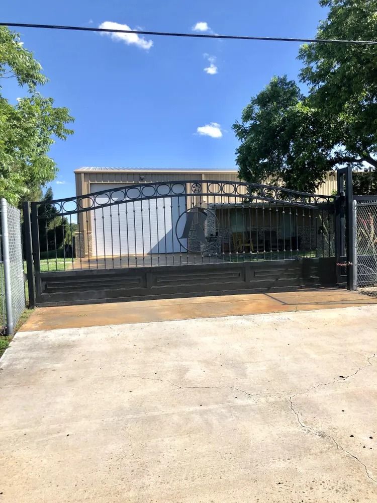 Black metal gate in front of a concrete driveway, leading to a warehouse. Blue sky with clouds.