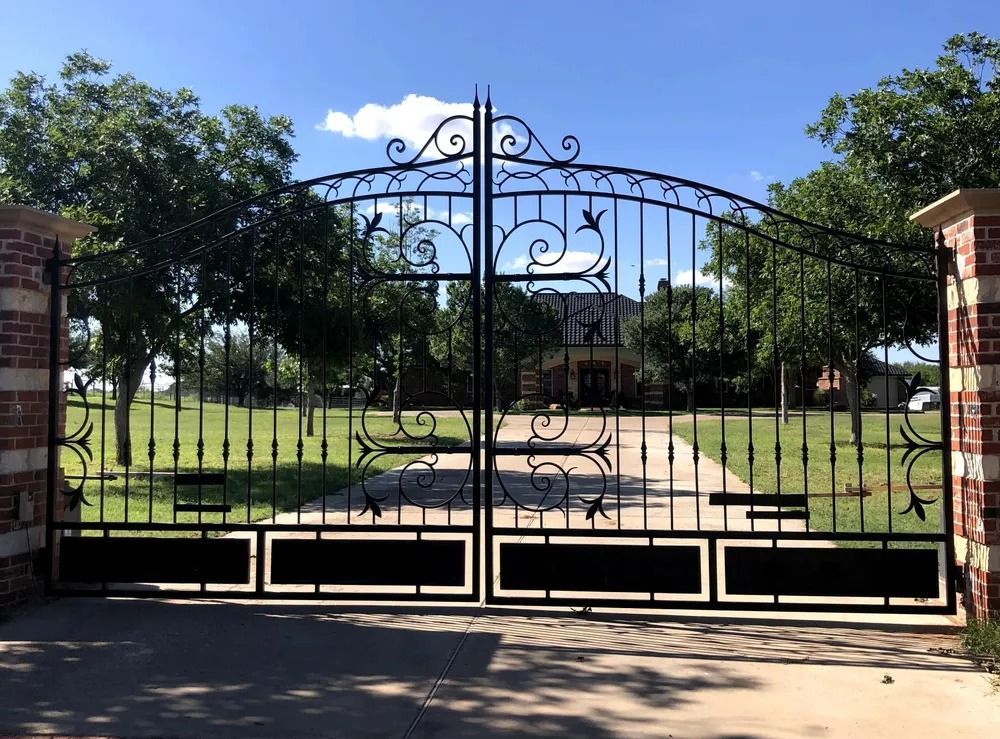 Black wrought-iron driveway gates with ornate designs, brick pillars, leading to a house, against a blue sky.