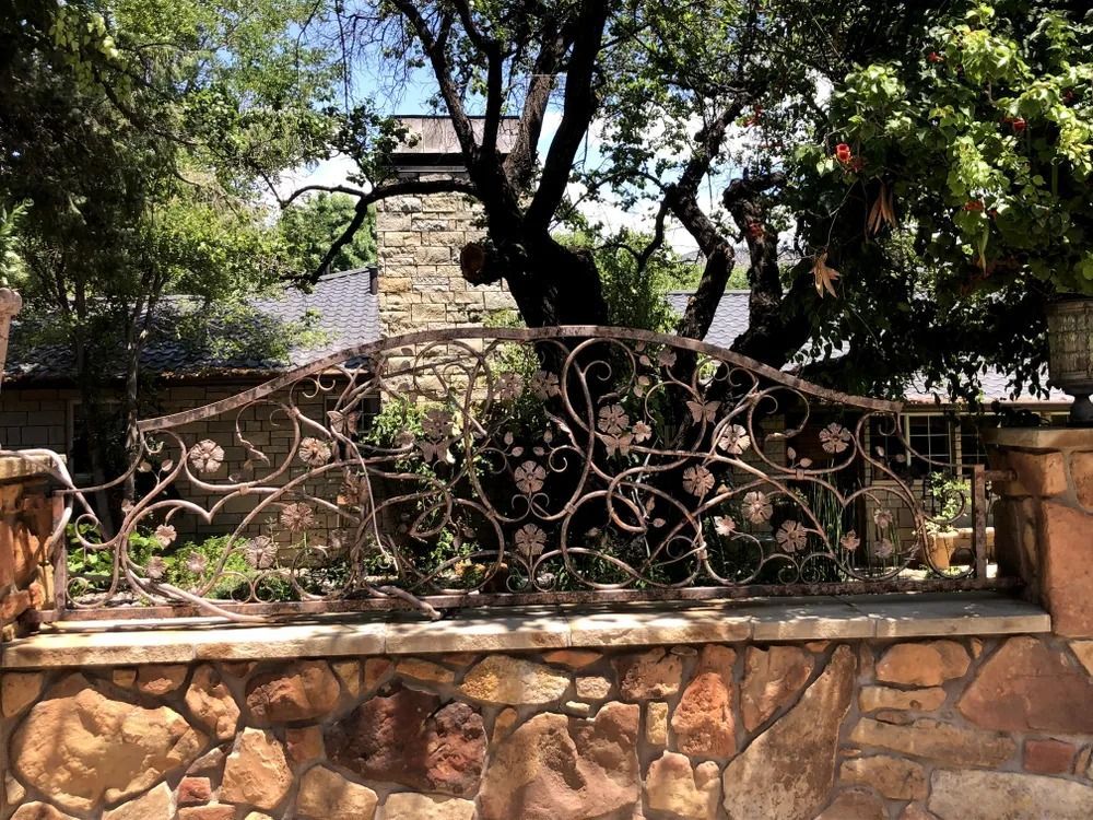 Wrought-iron fence with floral design atop a stone wall, in front of greenery and a building.