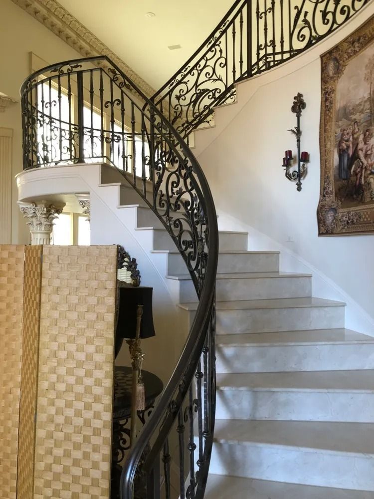 Elegant curving staircase with ornate wrought-iron railing, white steps, and a tapestry on the wall.