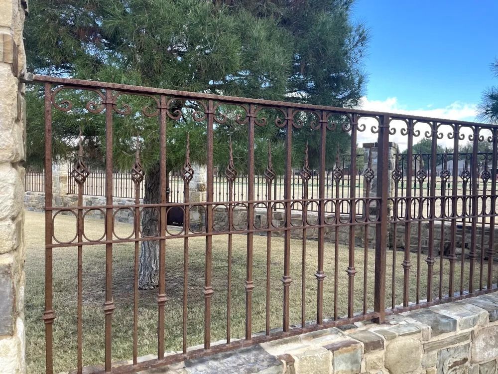 Rusty wrought iron fence atop a stone wall, enclosing a grassy area with trees and a blue sky.