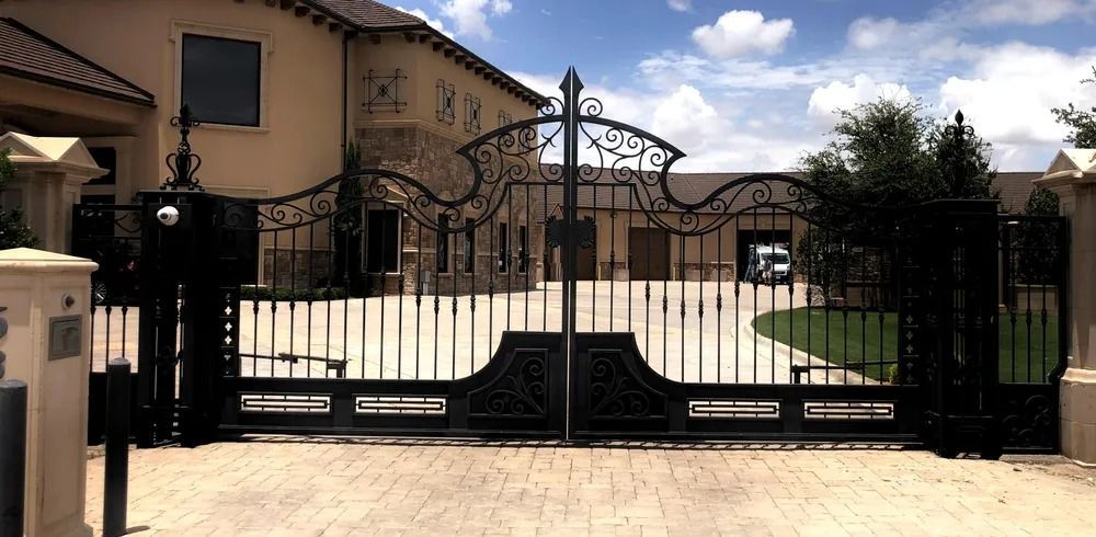 Ornate black iron gates in front of a large beige house with stone accents, on a brick driveway, under a partly cloudy sky.