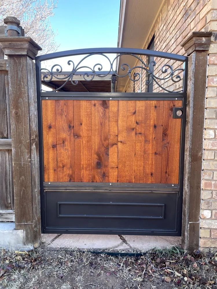 Wooden and metal gate with decorative ironwork, set between wooden posts and a brick wall.