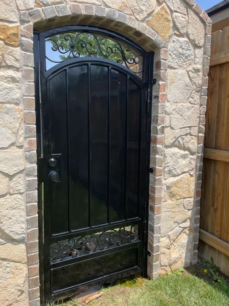 Black wrought iron gate set in a stone wall. Arch top has decorative scrollwork.