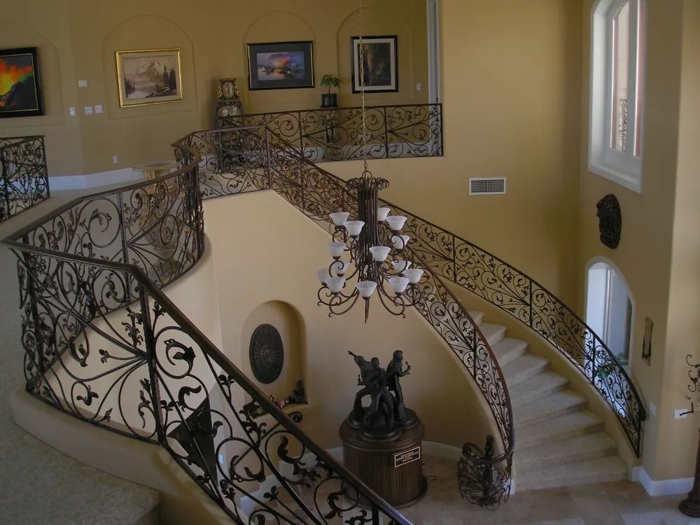 Two-story foyer with curved staircase, ornate wrought iron railing, chandelier, and artwork on cream-colored walls.
