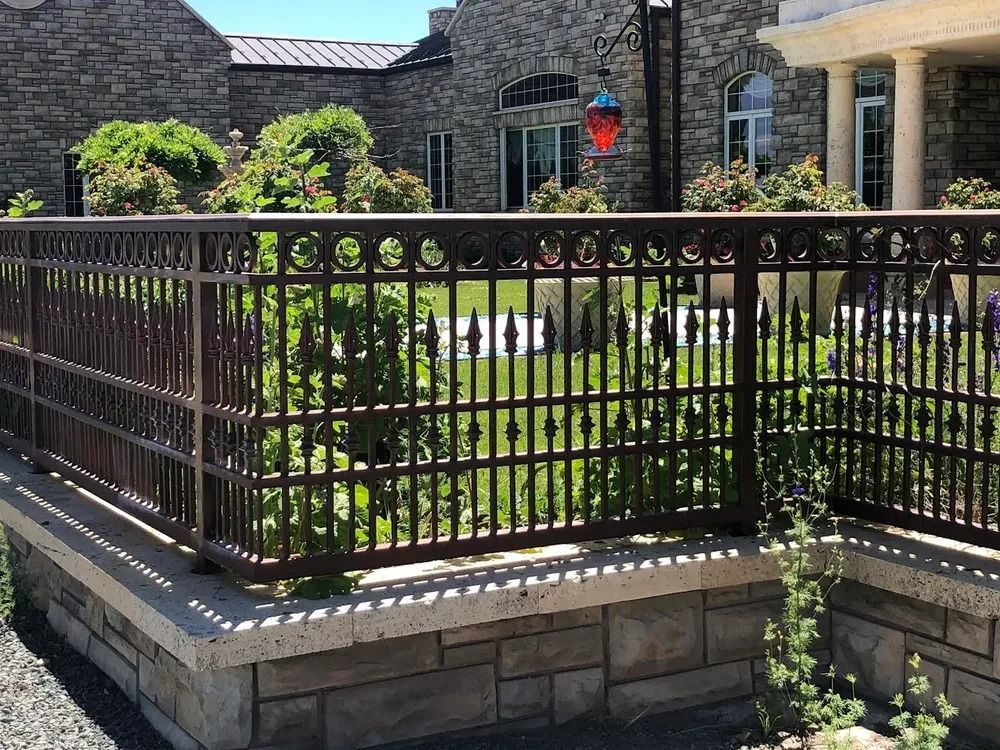 Brown wrought iron fence atop a stone retaining wall in front of a stone building with lush green foliage.