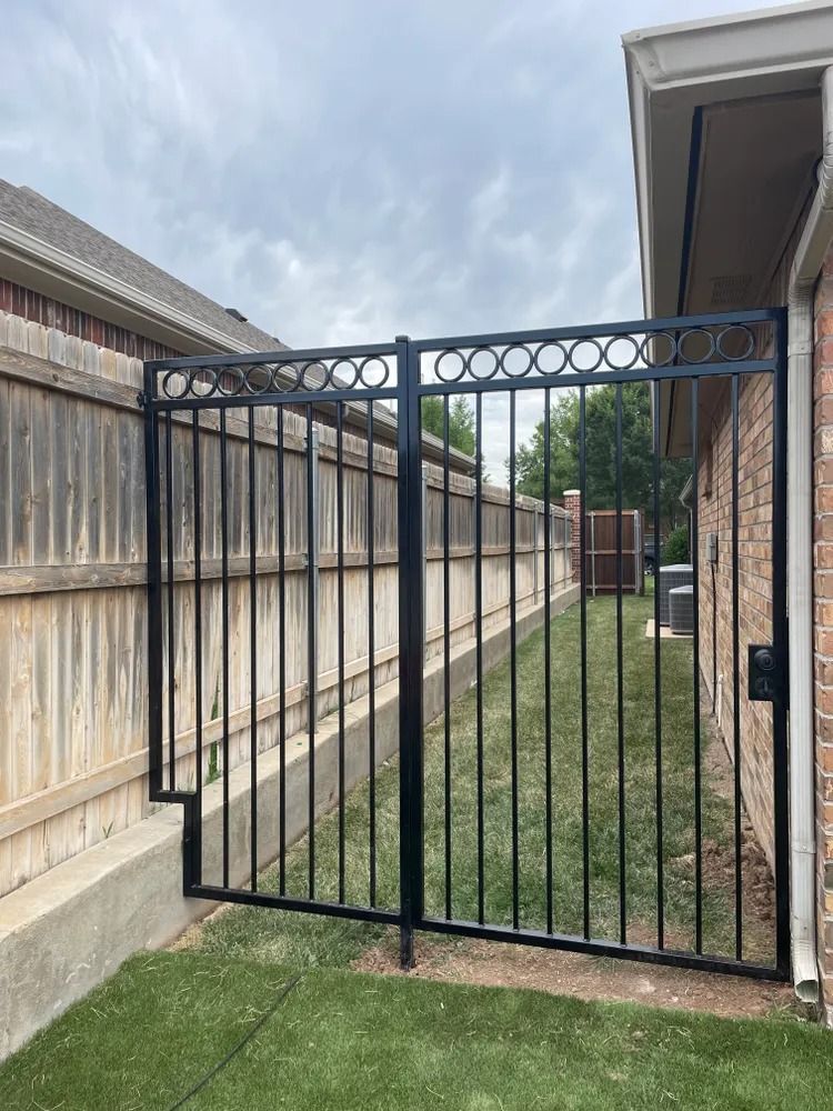 Black metal gate between wooden fence and brick wall, leading to a grassy yard.